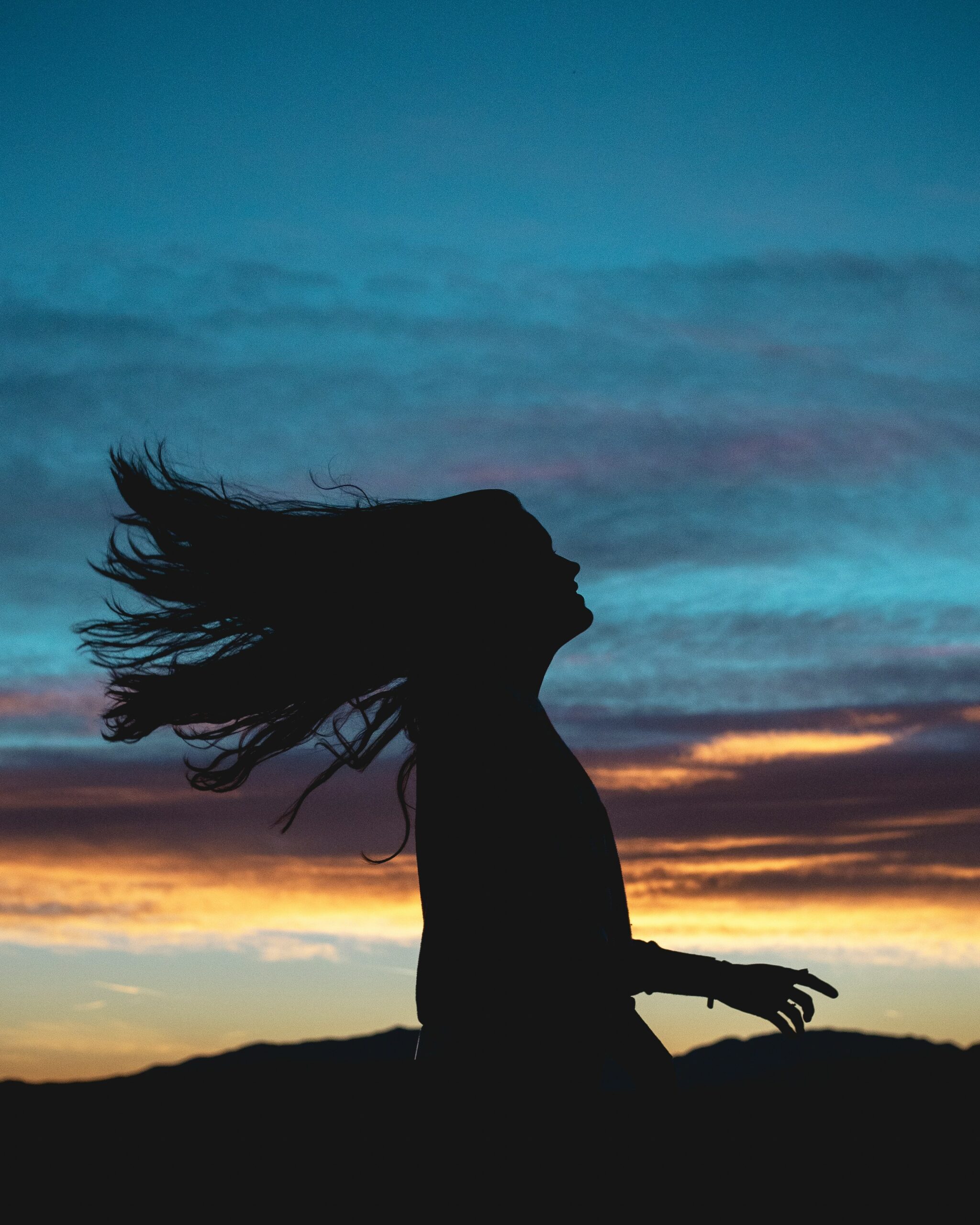 Silhouette of a woman with her hair flowing in the wind against a vivid sunset sky of blue, pink, and orange hues.